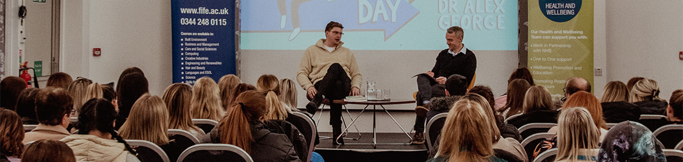 doctor alex george on stage with fife college staff member facing audience of students