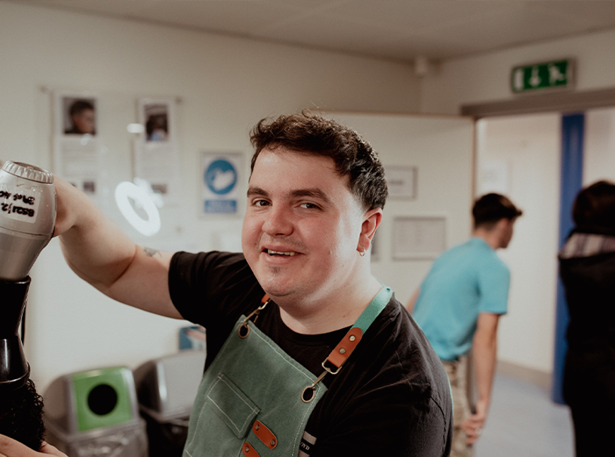 male barbering student smiling at camera holding hairdryer